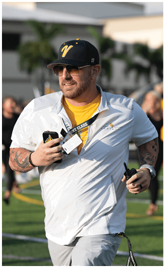 An adult wearing a black cap, sunglasses, a white jacket over a yellow shirt, and holding a walkie-talkie is outdoors on a grassy field with blurred people and buildings in the background. Descripci n generada con IA