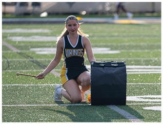 A person wearing a Vikings cheerleading uniform kneels on a sports field holding a drumstick next to a large black drum. Descripci n generada con IA