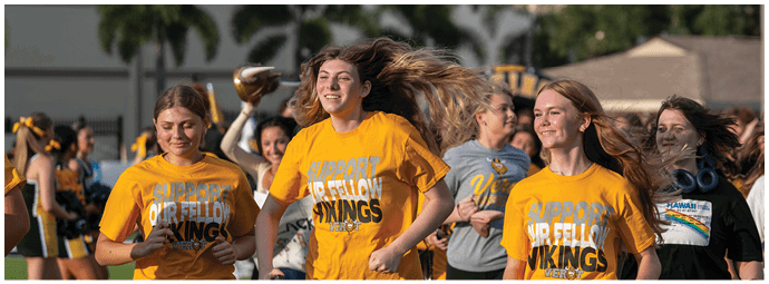 A group of people wearing yellow shirts with text are smiling and walking outdoors with palm trees and buildings in the background. Descripci n generada con IA