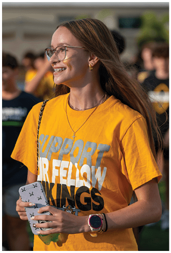 A person with long hair and glasses is smiling while holding a phone and wearing a bright yellow shirt with text supporting fellow Vikings at an outdoor event. Descripci n generada con IA