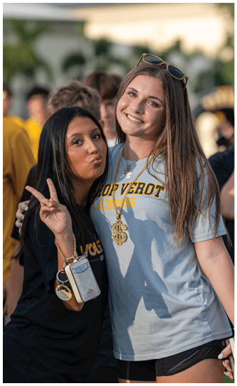 Two adults stand closely together smiling, one making a peace sign while wearing a black shirt and holding a phone, and the other wearing a light gray shirt with “Bishop Verot Lacrosse" text and sunglasses on their head, with several people in yellow shirts in the background. Descripci n generada con IA
