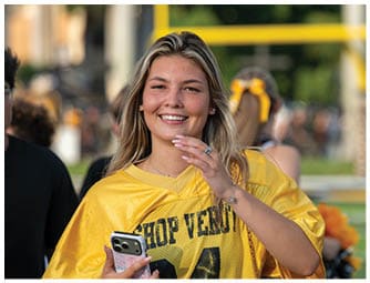 A person with long hair wearing a yellow sports jersey is smiling and holding a phone while standing outdoors with other people in the background. Descripci n generada con IA