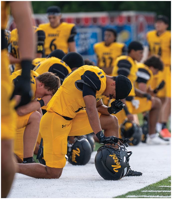 People wearing yellow and black sports uniforms are gathered closely together, some bending forward, likely preparing for or participating in a team activity on a field. Descripci n generada con IA
