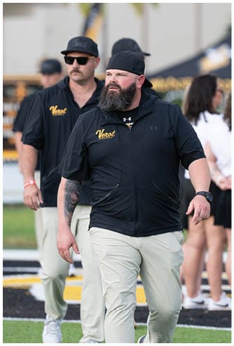 Two adults wearing black shirts with yellow text and light-colored pants walk on a sports field with people and tents in the background. Descripci n generada con IA