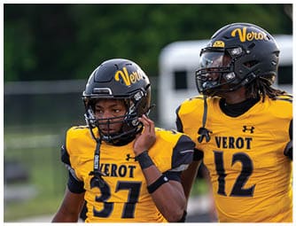 Two people wearing yellow and black football uniforms with helmets stand on a field, one adjusting their helmet strap. Descripci n generada con IA