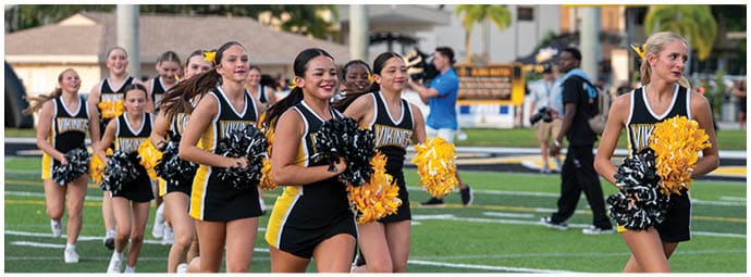 A group of adults in black and yellow cheerleading uniforms are smiling and holding pom-poms while standing on a sports field. Descripci n generada con IA