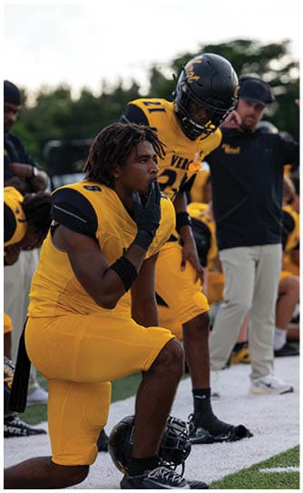 A person in yellow football gear kneels on one knee with a helmet on the ground while another person in similar gear and a coach stand nearby on the sidelines of a football field. Descripci n generada con IA