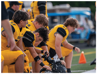 Four people in yellow and black sports uniforms are sitting on a bench on a field, with some holding helmets and looking focused. Descripci n generada con IA