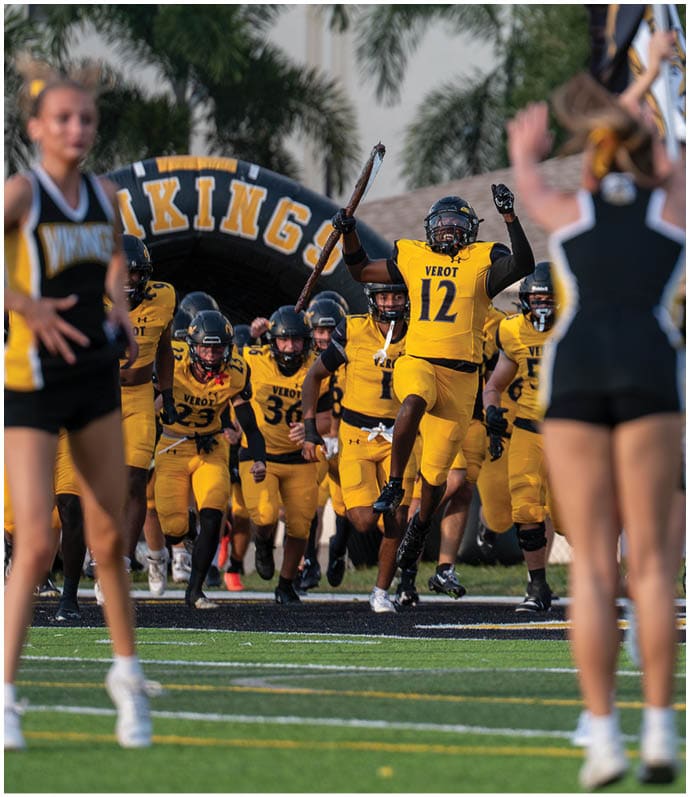 A group of athletes in yellow uniforms with “VEROT" on them are running onto a field through an inflatable tunnel labeled "VIKINGS" while cheerleaders in black and yellow outfits stand in the foreground. Descripci n generada con IA