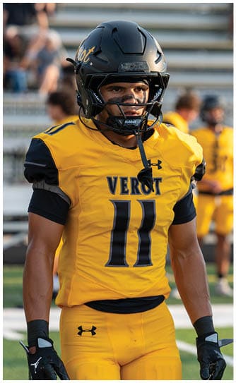 A person in a yellow football uniform with the number 11 and a black helmet stands on a field next to a referee in a black and white striped shirt with bleachers and other players blurred in the background. Descripci n generada con IA