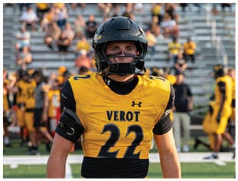 A person wearing a yellow and black football uniform with the number 22 and the word VEROT on the chest stands on a field with blurred spectators and other players in the background. Descripci n generada con IA