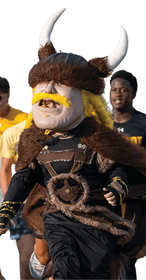A person dressed in a Viking mascot costume is running on a field followed by a group of people wearing casual sportswear. Descripci n generada con IA