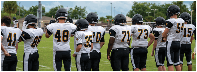Four people wearing football helmets and white jerseys with “Bishop Verot" written on them stand side by side on a sports field with empty bleachers in the background. Descripci n generada con IA