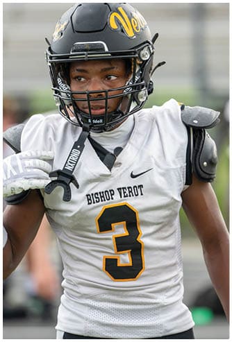 A person wearing a black and yellow Bishop Verot sports jersey stands with arms crossed and a thoughtful expression while resting their chin on one hand. Descripci n generada con IA