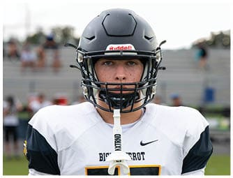 A person wearing a black football helmet and a white and black football jersey is smiling on a sports field with empty bleachers in the background. Descripci n generada con IA