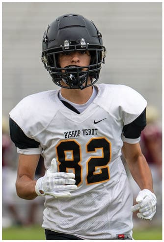 A person wearing a black helmet and a white football uniform with “Bishop Verot" on the jersey is running on a field with another player blurred in the background. Descripci n generada con IA