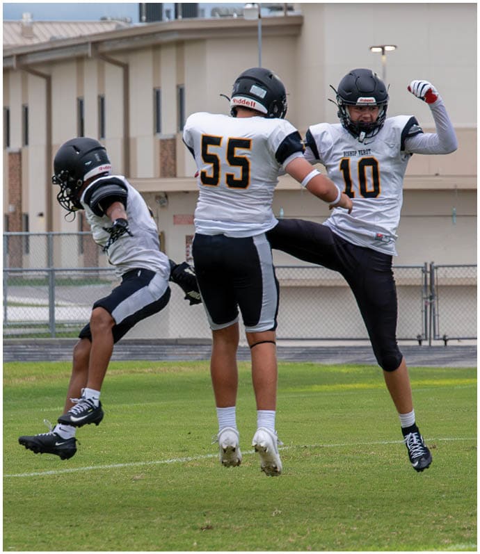 Three people wearing white football uniforms with helmets walk together on a green field during a game or practice. Descripci n generada con IA