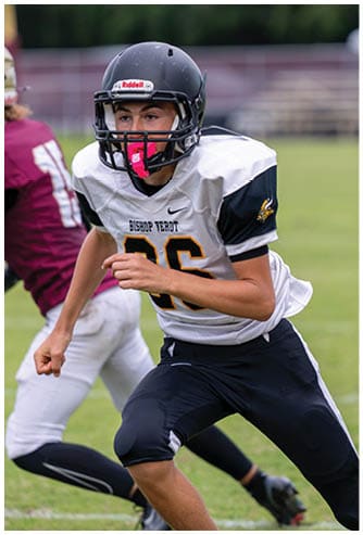 A person wearing a black football helmet and white Bishop Verot jersey with the number 66 stands on a football field with blurred players and bleachers in the background. Descripci n generada con IA