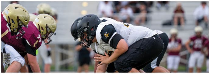 A group of people wearing football helmets and white jerseys with black and gold numbers are gathered closely on a field. Descripci n generada con IA