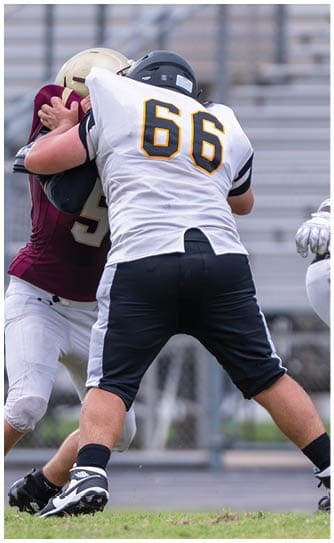 A person wearing a football uniform is kicking a ball on a grassy field with one leg raised high and the other planted on the ground. Descripci n generada con IA