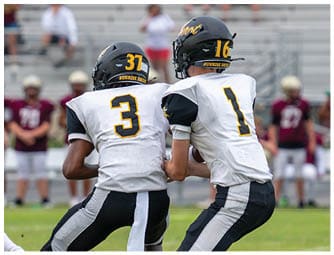Two people in football uniforms are engaged in a tackle on a field while another player in uniform runs nearby with spectators in the background. Descripci n generada con IA
