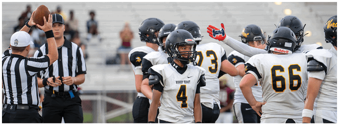 People in football uniforms are actively engaged in a play on a field with bleachers and spectators in the background. Descripci n generada con IA