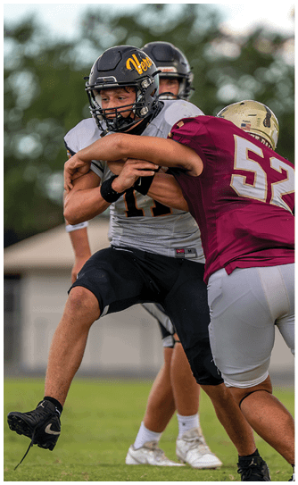People wearing white football uniforms with numbers 12 and 18 stand on a field holding helmets while facing away from the camera. Descripci n generada con IA