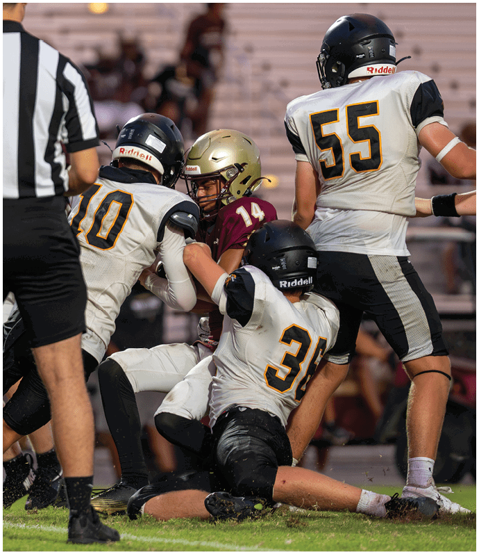 Two people in white sports uniforms with black helmets are running on a football field while two others in green uniforms are nearby. Descripci n generada con IA