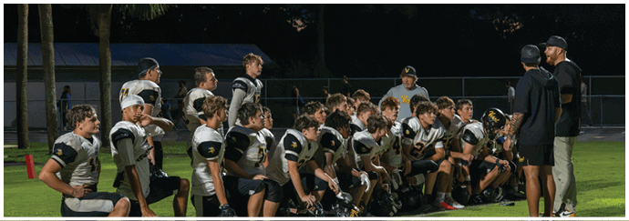 A group of people in football uniforms are kneeling on a field at night while two people stand nearby, one speaking into a microphone. Descripci n generada con IA