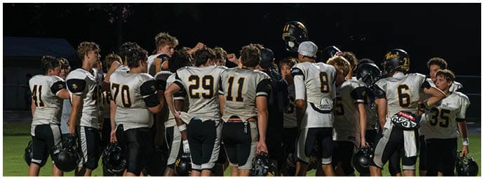 A group of people in sports uniforms and casual clothing are gathered on the sidelines of a football field, some standing and some sitting, with a goalpost and bleachers in the background. Descripci n generada con IA