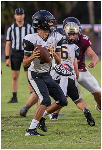 Two people in football uniforms are engaged in a play on a field, with one holding the ball and the other preparing to receive or block. Descripci n generada con IA