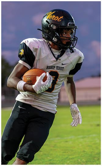 People in football uniforms are actively engaged in a nighttime game on the field with one player in white attempting to block or reach over players in green. Descripci n generada con IA