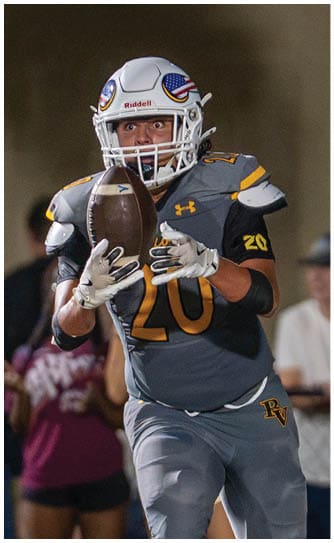 A person in a yellow football uniform is running with the ball while another person in a white football uniform is reaching out to try to tackle them on a lit football field. Descripci n generada con IA