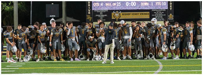 A group of people wearing football uniforms stand closely together on a field at night with stadium lights in the background. Descripci n generada con IA