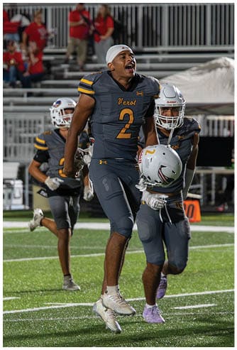 A person wearing a yellow and black football uniform with the number 2 is on a football field at night, raising their hand with a thumbs-up gesture. Descripci n generada con IA