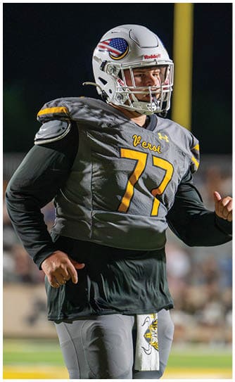 A person in a yellow sports uniform is facing another person wearing a black cap and black jacket, who is smiling on a sports field. Descripci n generada con IA