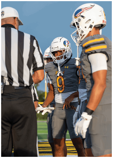 A person in a yellow sports uniform is running with a football while two people in white uniforms are attempting to tackle them on a football field. Descripci n generada con IA