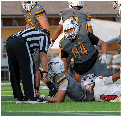 Four football players in yellow uniforms and one player in a white uniform are positioned on a field during a game with a crowd and cheerleaders in the background. Descripci n generada con IA