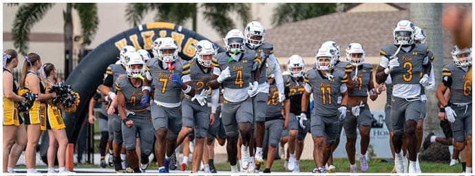 People in yellow and white football uniforms are engaged in a play on a football field with a goalpost and bleachers in the background. Descripci n generada con IA