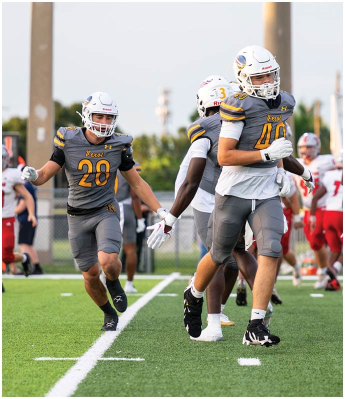 A person in a yellow football uniform is running on a field while holding a football, with teammates and coaches in similar uniforms and attire cheering in the background. Descripci n generada con IA