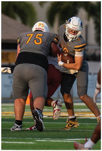 Two people in football uniforms are engaged in a close physical play, with one wearing a yellow and black uniform and the other in a white and maroon uniform. Descripci n generada con IA