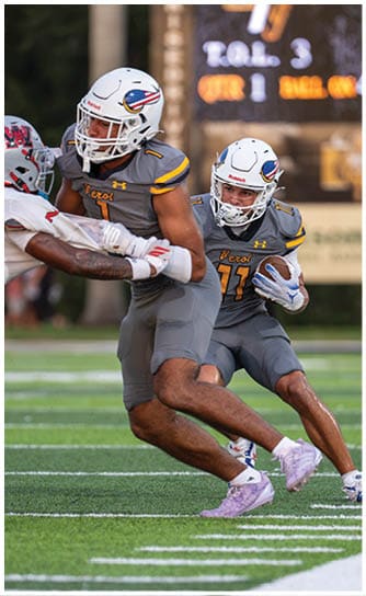 Two adults in yellow football uniforms with black helmets are on a field, one holding a football and the other standing nearby. Descripci n generada con IA