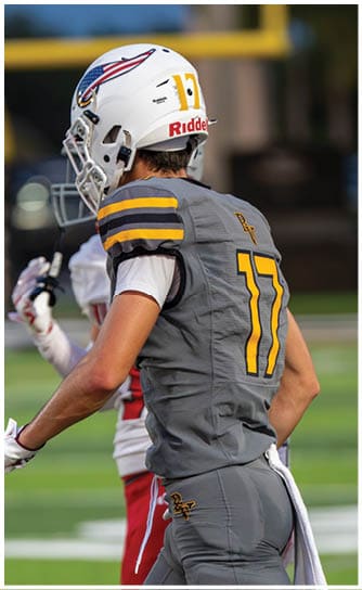 A person in a yellow football uniform with the number 17 and the word Verot is sitting on the sideline reaching out while another person in casual clothing stands nearby. Descripci n generada con IA