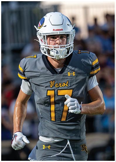 A person in yellow sports gear is in the foreground while a person in white sports gear with a gold helmet is in the background on a field with spectators behind them. Descripci n generada con IA