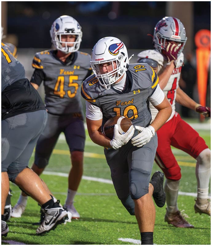 A person in a yellow football uniform with the number 20 is on the right side of the image facing a person in a white football uniform with the number 8 and a gold helmet, with a blurred crowd in the background. Descripci n generada con IA