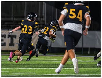 Three people in black and yellow football uniforms are running on a field during a game at night. Descripci n generada con IA