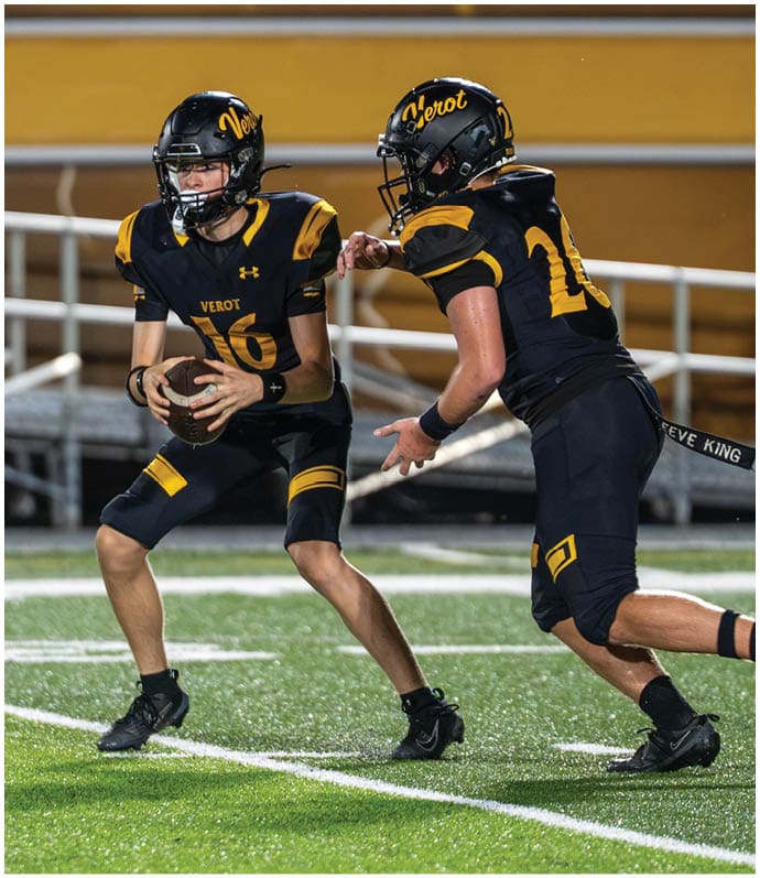Two people in black and yellow football uniforms with helmets are on a field, with one holding a football and preparing to pass while the other reaches out. Descripci n generada con IA