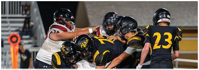 People wearing helmets and football uniforms are engaged in a close physical play on a football field at night. Descripci n generada con IA