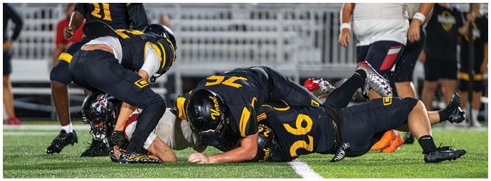 Three people in football uniforms are on the ground during a play on a green field with white yard lines. Descripci n generada con IA