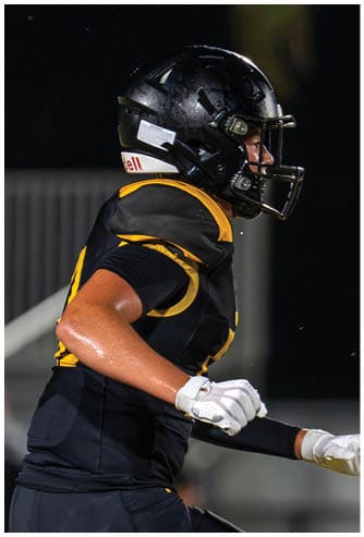 A person wearing a black and yellow football uniform and helmet is running on a field at night. Descripci n generada con IA